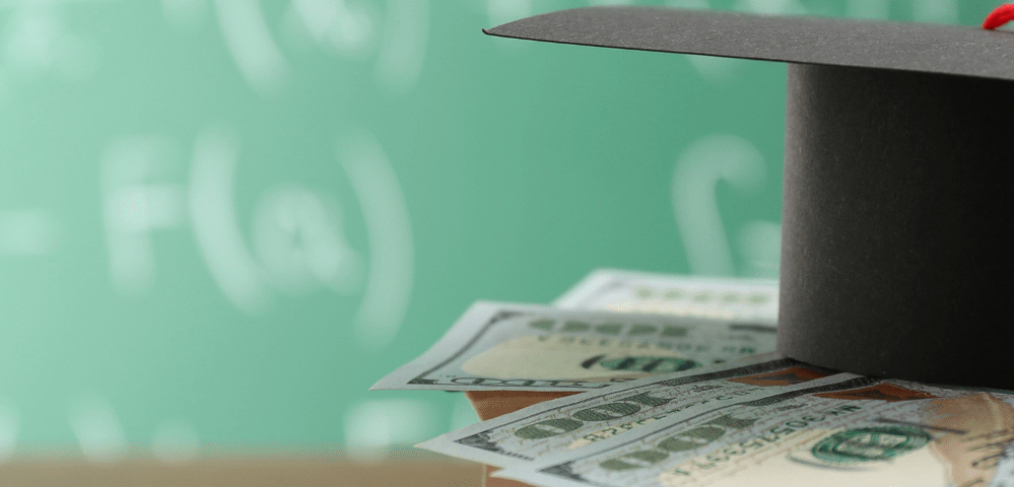 Grad cap on sits on top of $100 dollar bills, with green chalboard in background with equations written on the board