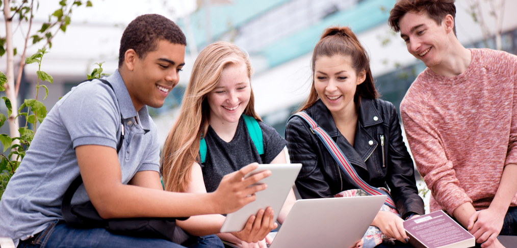 4 students smiling, sitting outside, looking at a tablet and laptop