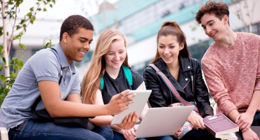 4 students smiling, sitting outside, looking at a tablet and laptop