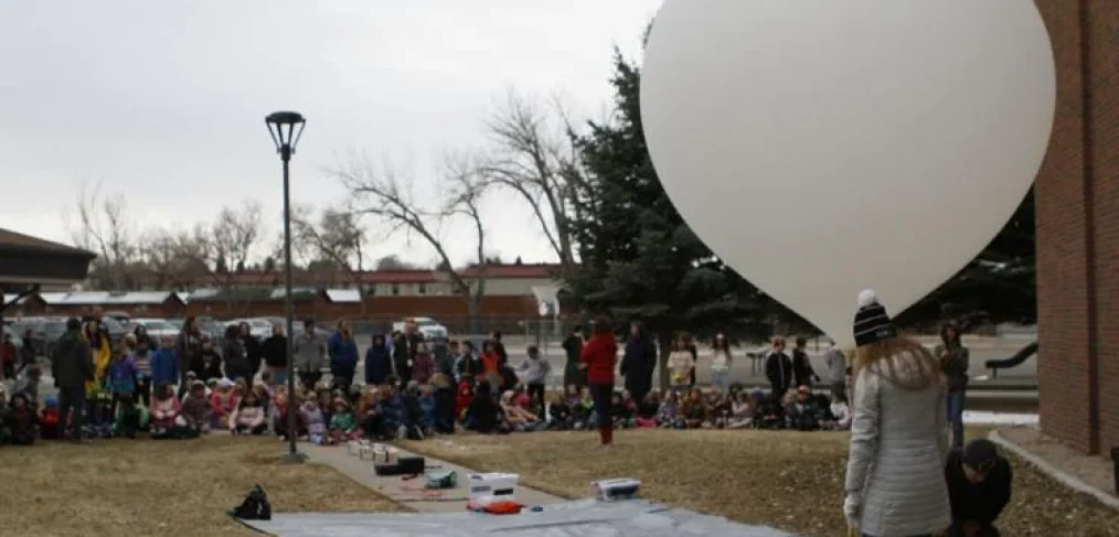 Elementary school kids and teachers sit outside watching the preparations for a balloon launch with payloads laid out on a tarp and another educator holding the balloon.