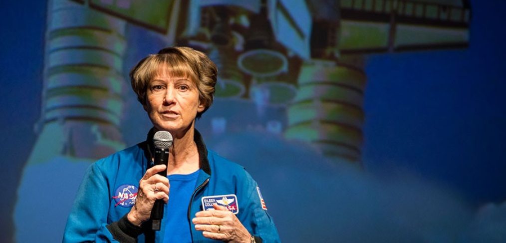 A person wearing a blue NASA flight jacket stands on a stage holding a handheld microphone. A large projected image of a space shuttle with visible engines appears in the background behind the speaker.