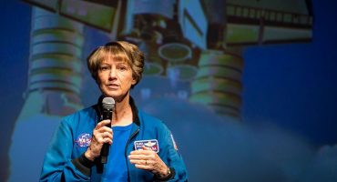 A person wearing a blue NASA flight jacket stands on a stage holding a handheld microphone. A large projected image of a space shuttle with visible engines appears in the background behind the speaker.